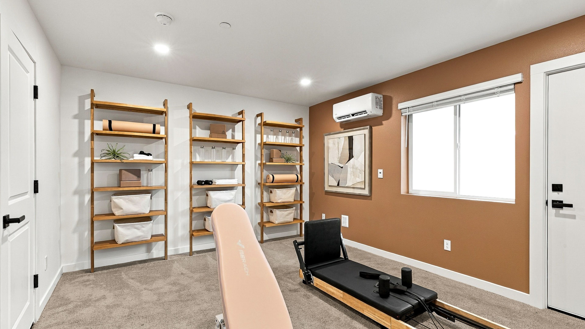 Home fitness room with Pilates-style reformer and exercise bench, three open wood shelving units with fitness accessories, orange accent wall, window, artwork, and mini-split unit.