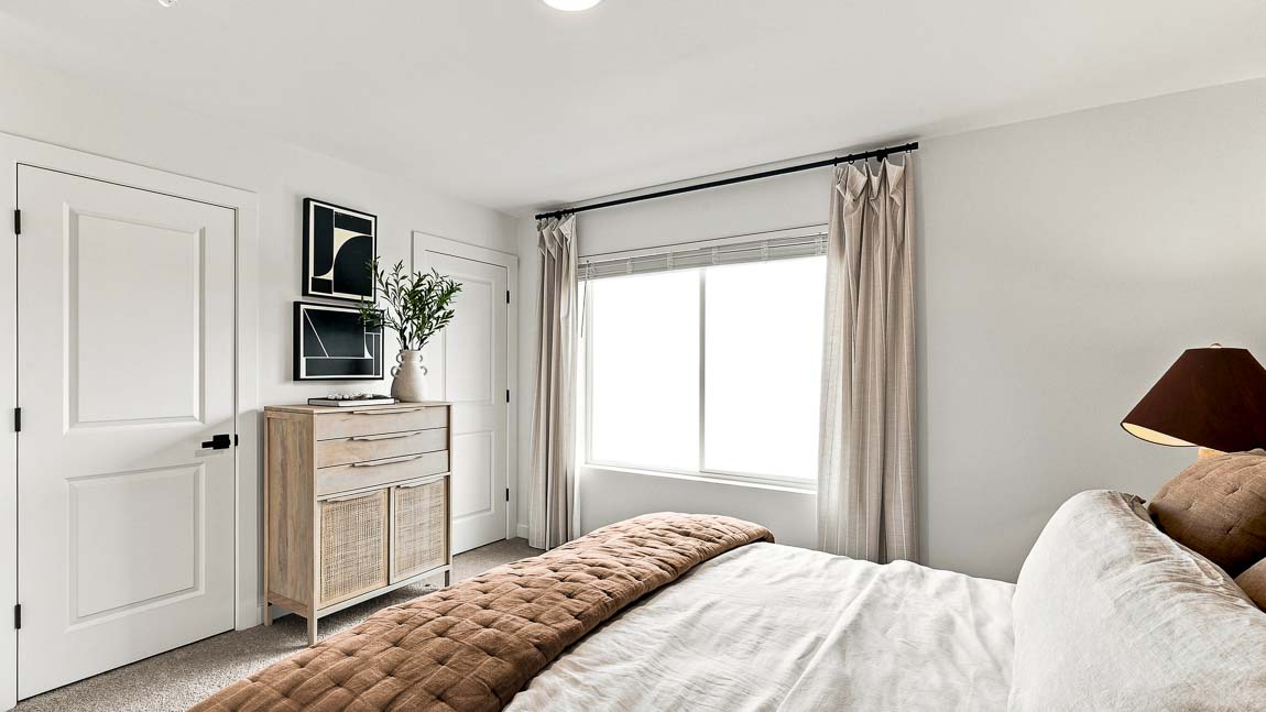 Bedroom viewed across the bed toward a large curtained window, with light carpet, wood dresser, framed black-and-white artwork, plant, and white interior doors.