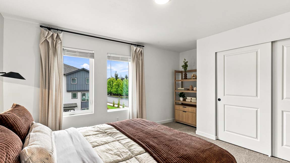 Bedroom with white and brown bedding in the foreground, two tall windows with beige curtains, wood storage cabinet with plant and artwork, and white closet doors.