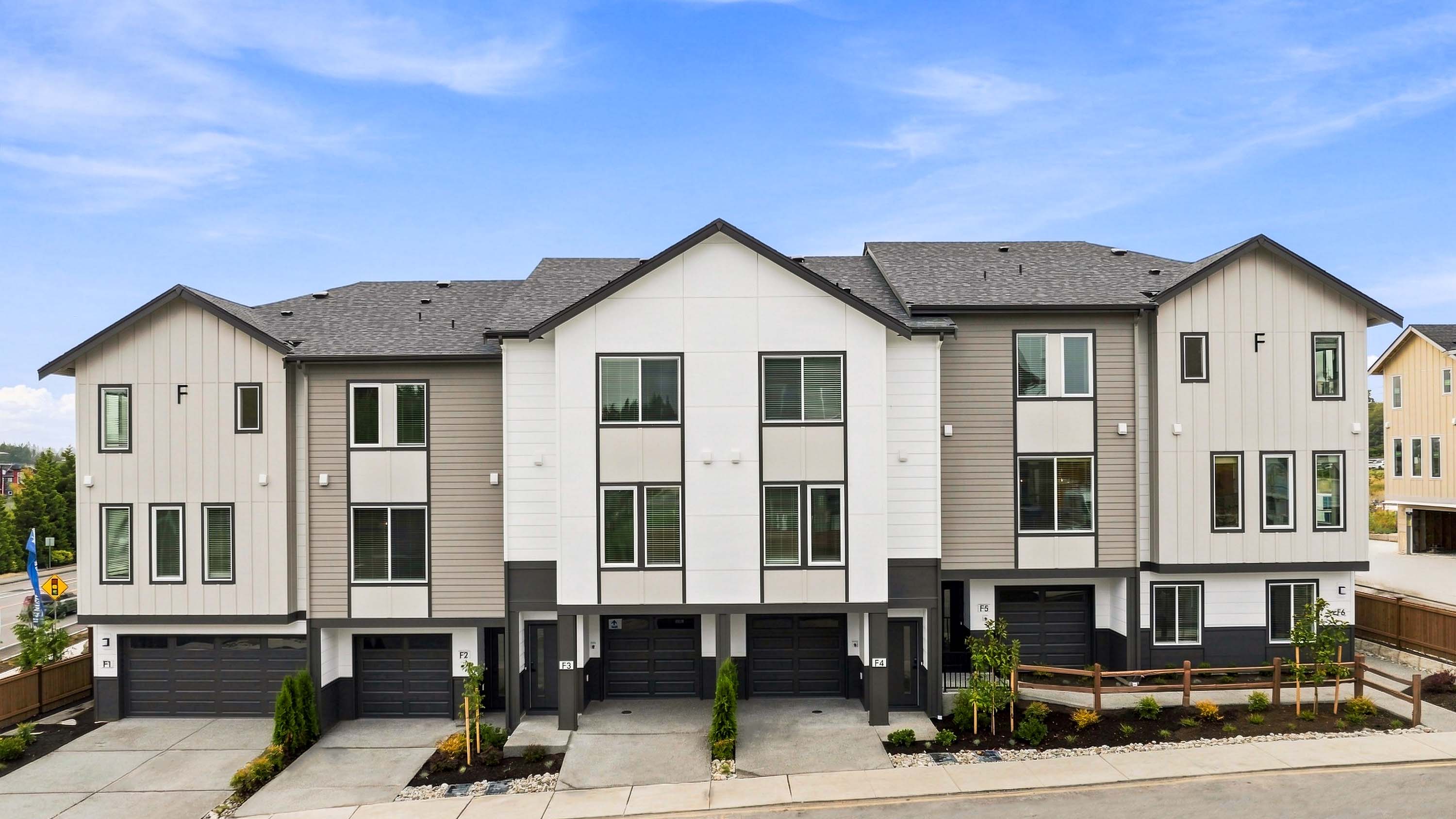 Exterior view of a row of modern three-story townhomes with white and gray siding, dark garage doors, multiple windows, peaked rooflines, landscaping, and front driveways.