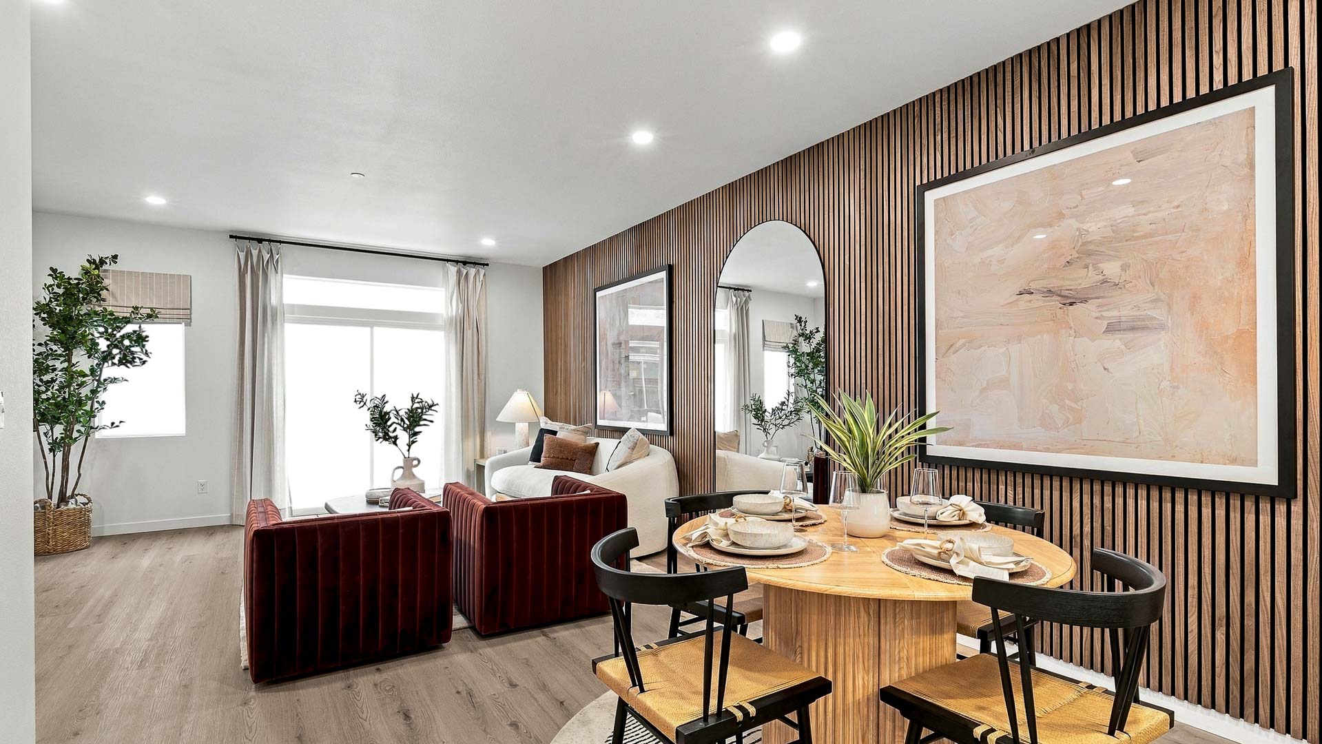 Open dining and living area with a round four-seat table, wood slat accent wall, framed artwork and arched mirror, white sofa, rust-colored chairs, and large windows.