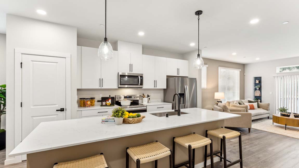 Wide view of a modern kitchen with light wood cabinets, stainless steel appliances, a long white island with four brown stools, and a dining area beyond.