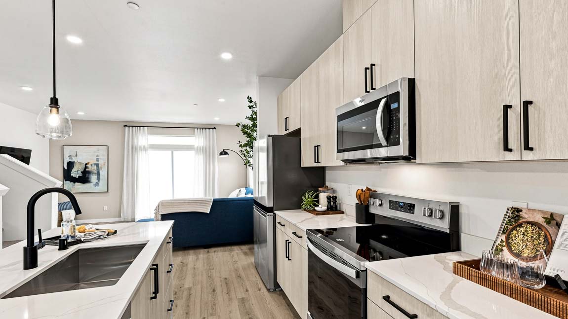 Modern galley-style kitchen with light wood cabinetry, quartz countertops, stainless steel appliances, pendant lighting, and an open-concept layout connecting to the living area.