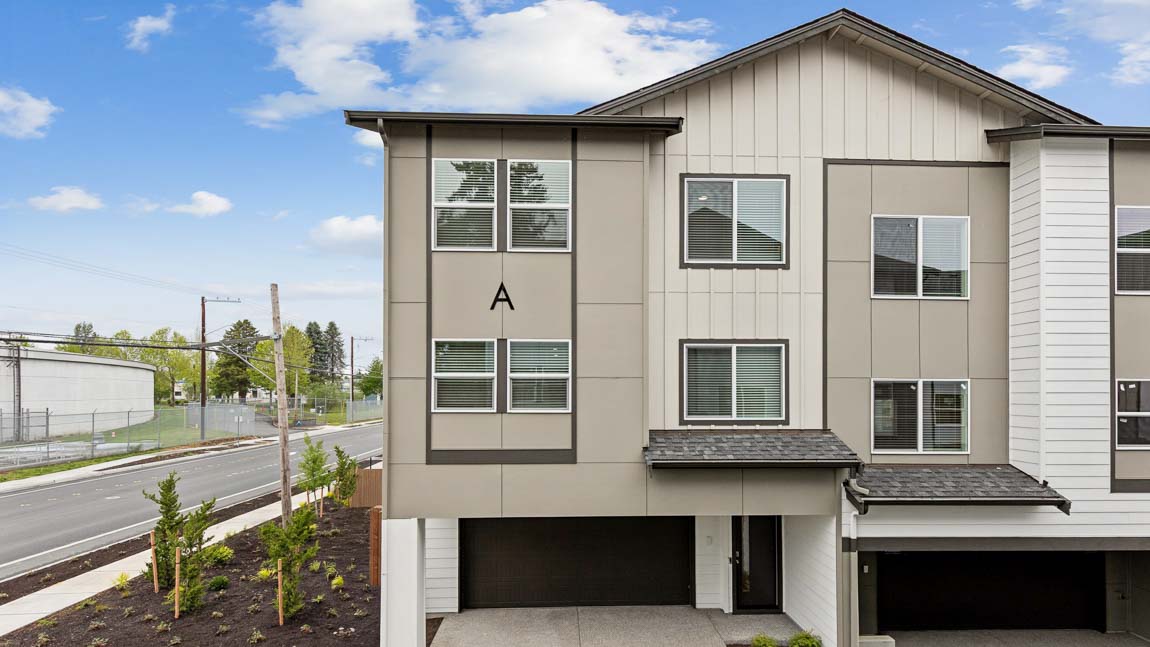 Front exterior of a three-story end-unit townhome featuring gray panel siding, attached garage, covered entry, and landscaped surroundings.