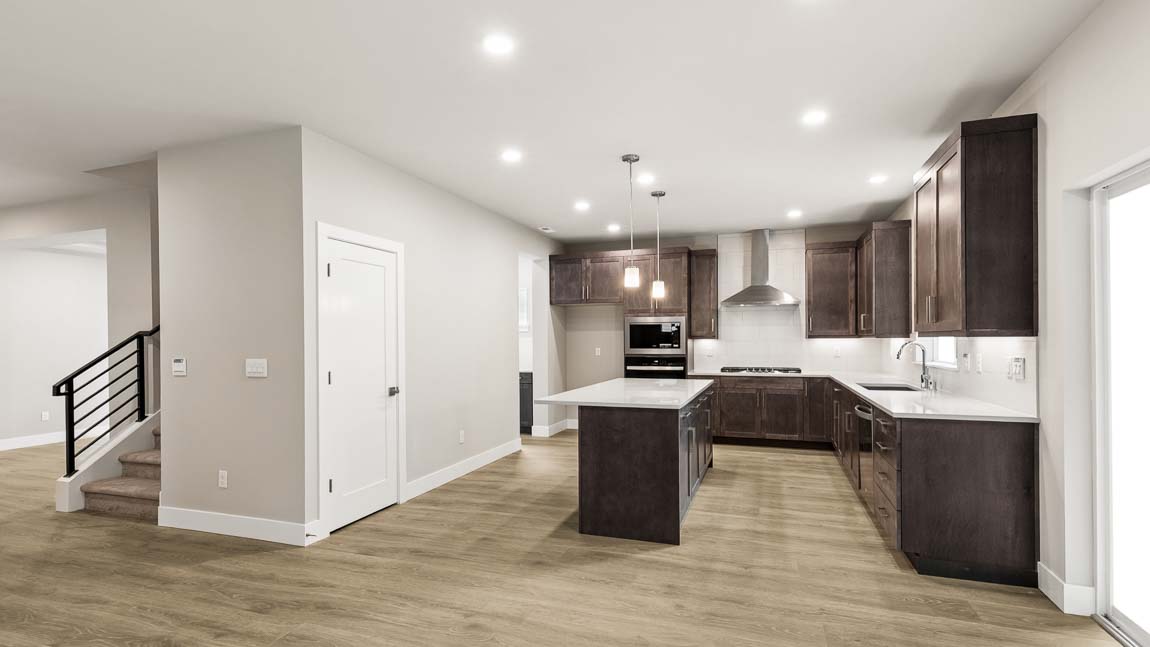 Bright gourmet kitchen featuring a large quartz island, dark wood cabinetry, pendant lighting, and open living space with multiple windows.