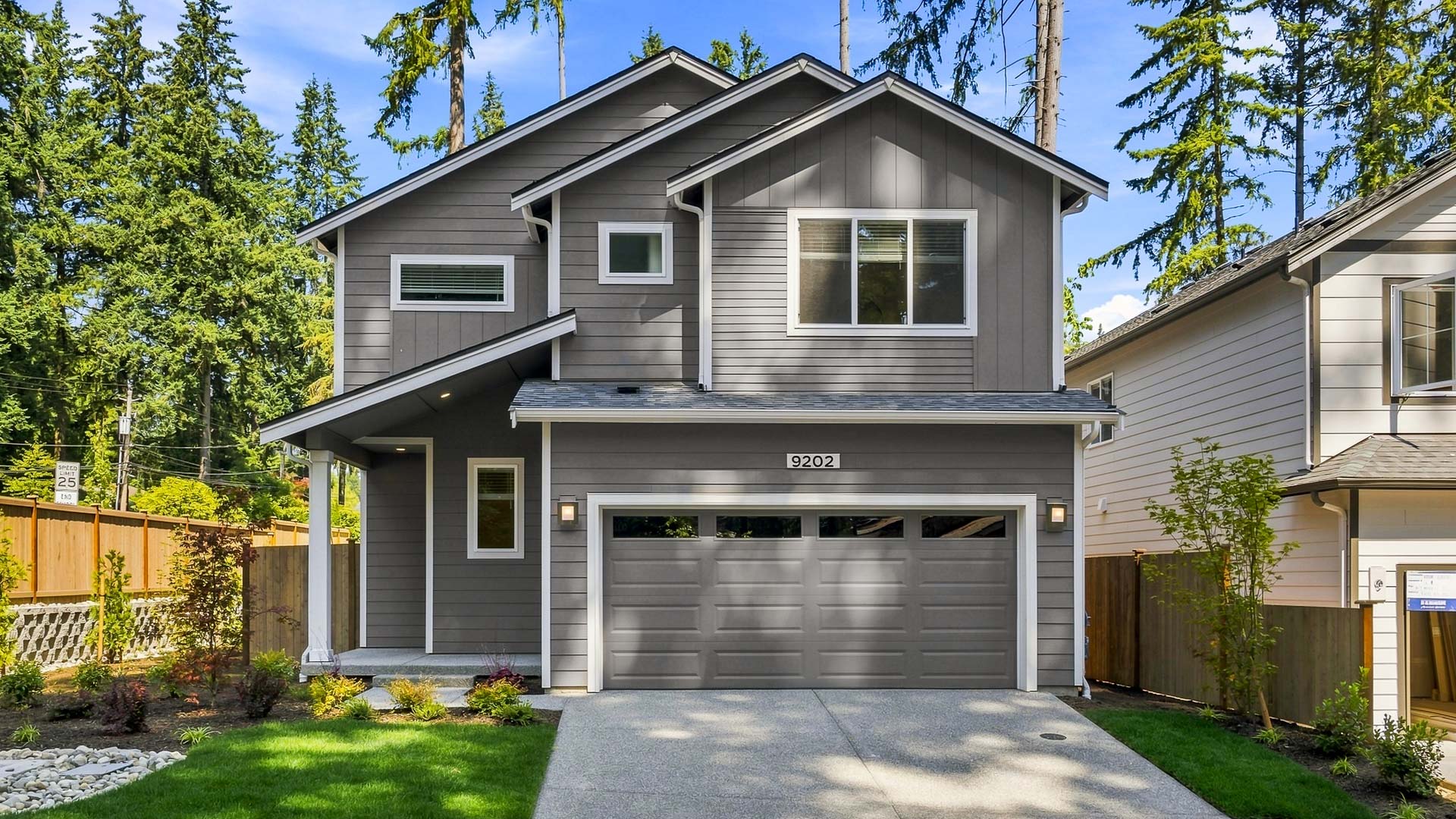 Two-story gray craftsman-style home with attached two-car garage, covered front entry, landscaped yard, and tall evergreen trees surrounding the property.