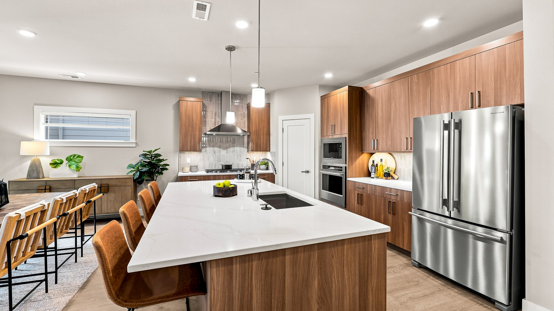 Modern kitchen with large white waterfall island, brown barstools, wood cabinetry, stainless steel appliances, and adjacent dining area.