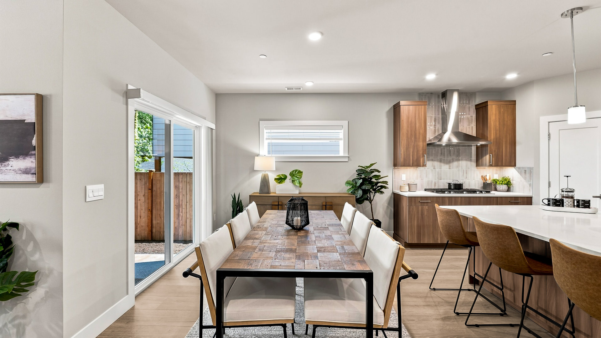 Dining room with long rectangular table, upholstered chairs, large window, and view into the kitchen.