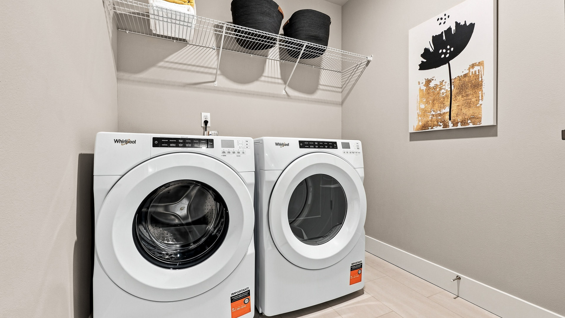 Laundry room with front-loading washer and dryer, overhead shelving, and wall-mounted artwork.