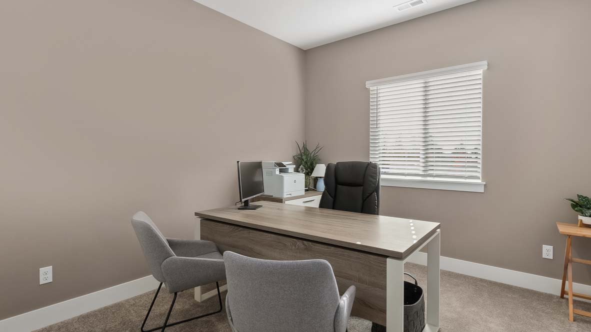 Home office with desk, guest seating, large window, and neutral-toned walls.