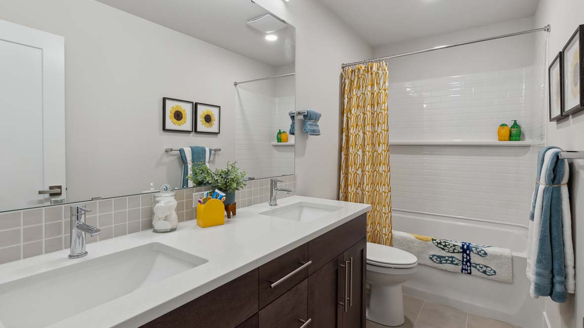 Bathroom with double-sink vanity, large wall mirror, white countertop, toilet, and bathtub with patterned shower curtain.