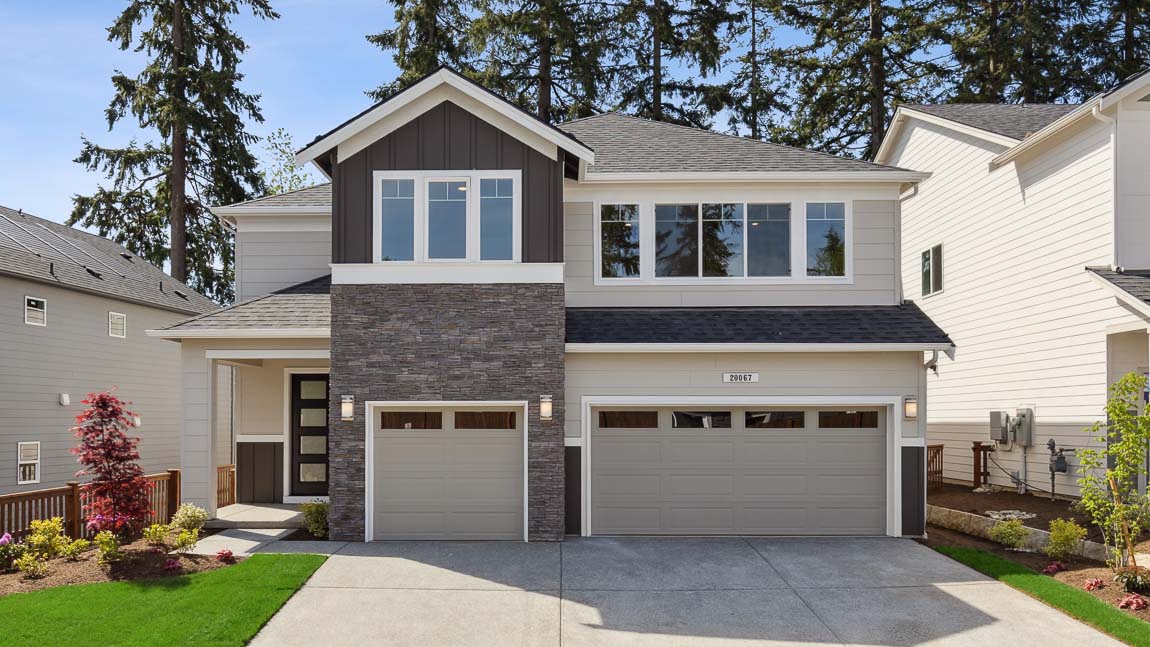 Two-story craftsman-style home with stone veneer entry feature, attached three-car garage, landscaped front yard, and mature trees in the background.