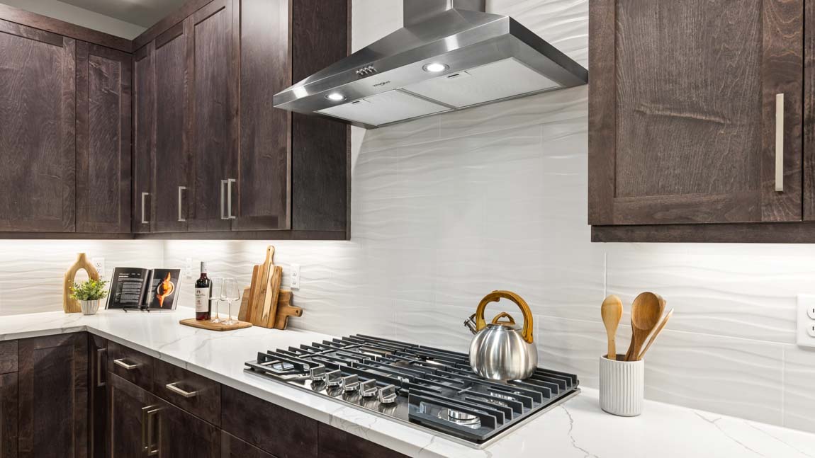Kitchen close-up with quartz island countertop, gas cooktop, stainless steel range hood, and dark wood cabinets.