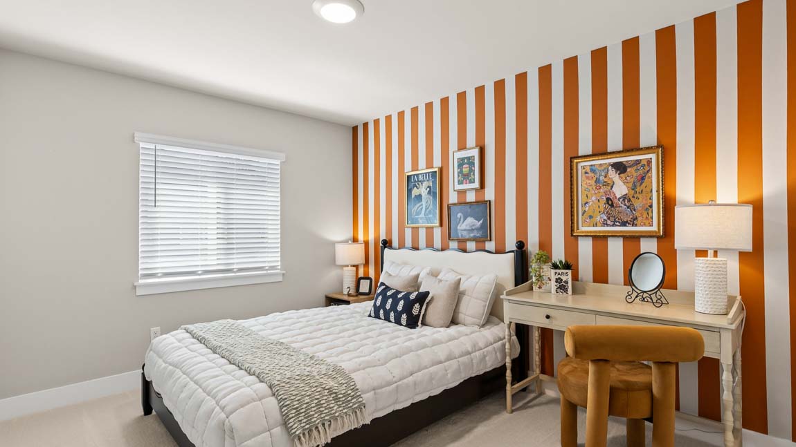 Secondary bedroom with white bedding, orange-and-white striped accent wall, writing desk, upholstered chair, and framed artwork.