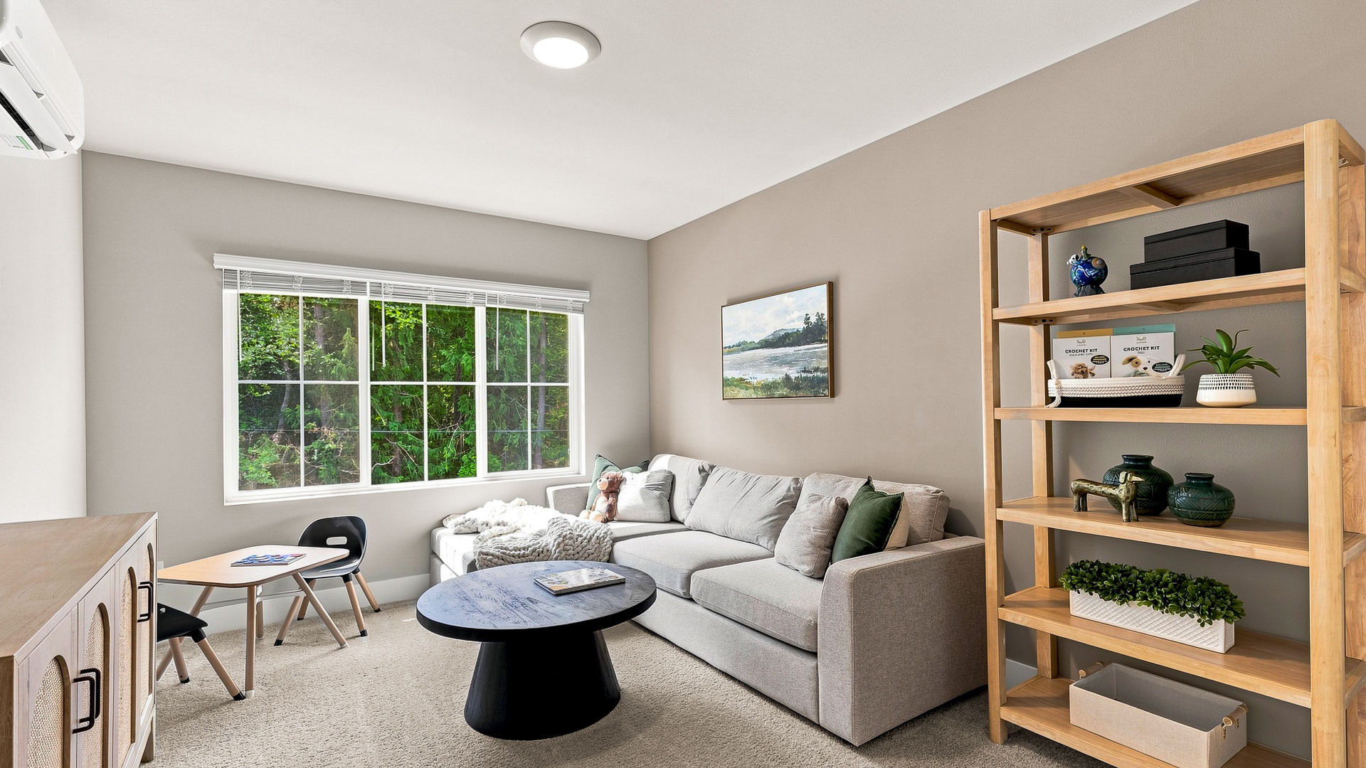 Bonus room with a gray sectional, round black coffee table, children's activity table, wood shelving, and a large window.
