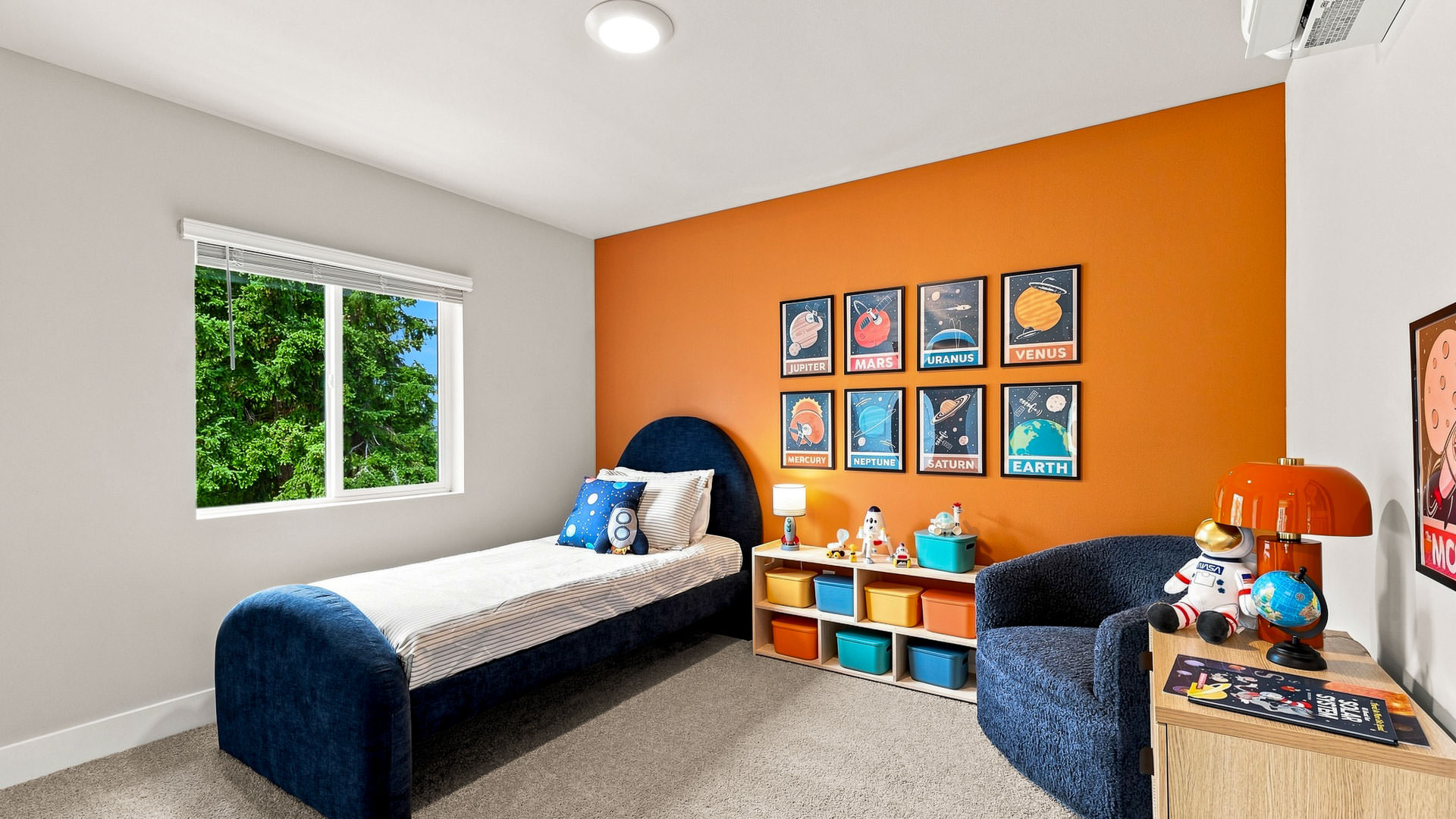Space-themed bedroom with an orange accent wall, navy upholstered bed, planet posters, toy storage, and a blue chair.