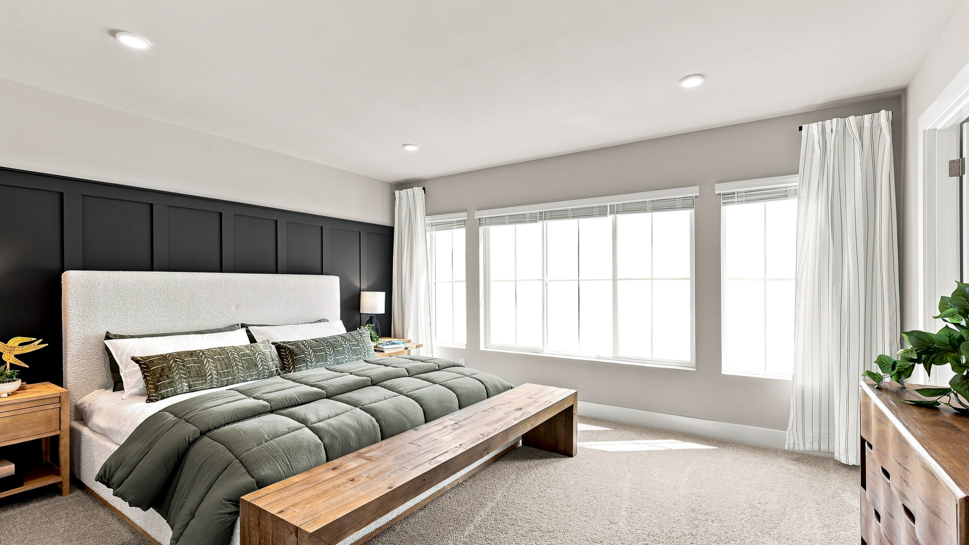 Primary bedroom with green bedding, upholstered headboard, black paneled accent wall, wood bench, and large windows.