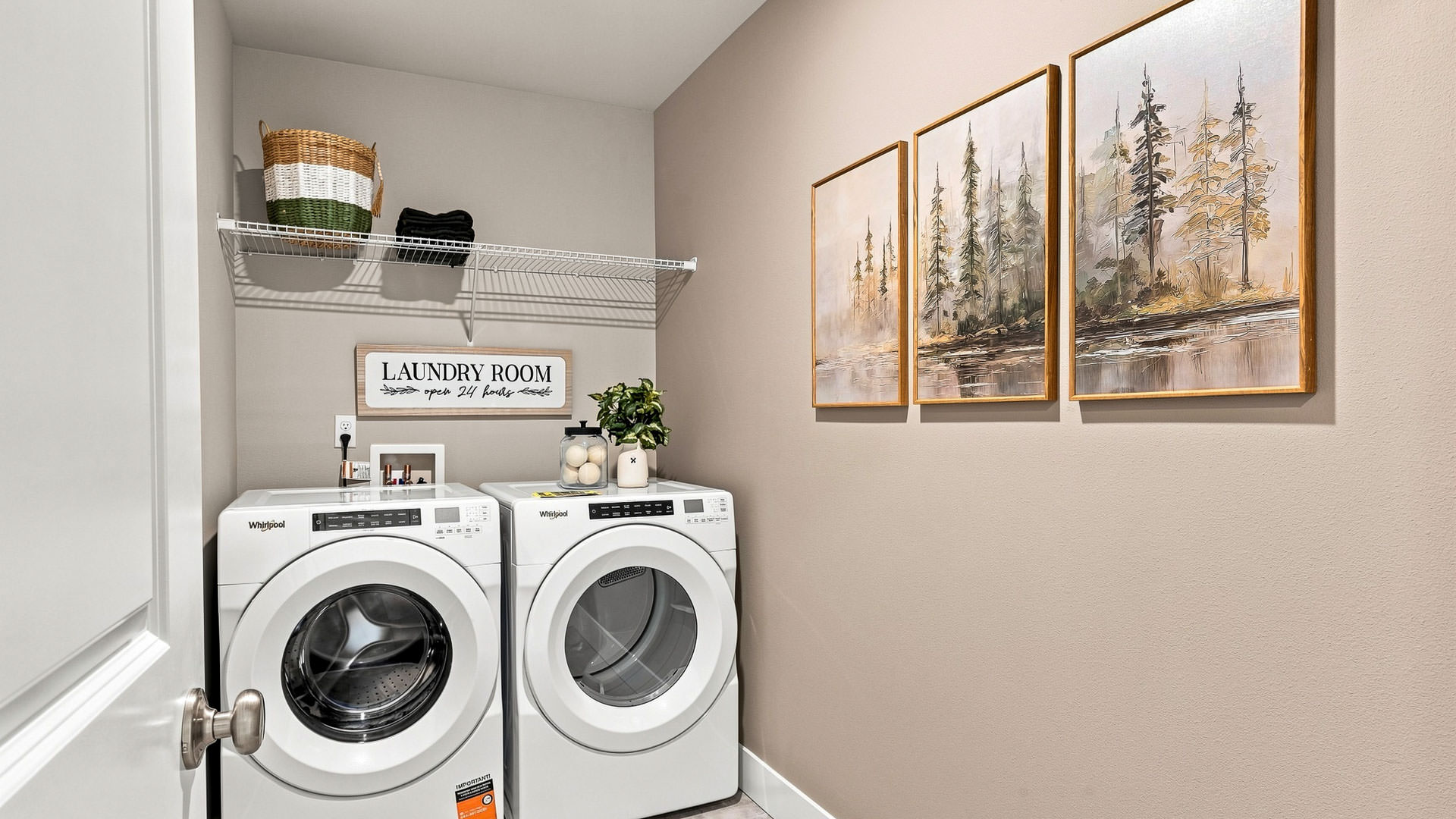 Laundry room with side-by-side white washer and dryer, wire shelf, storage basket, and three framed forest prints.