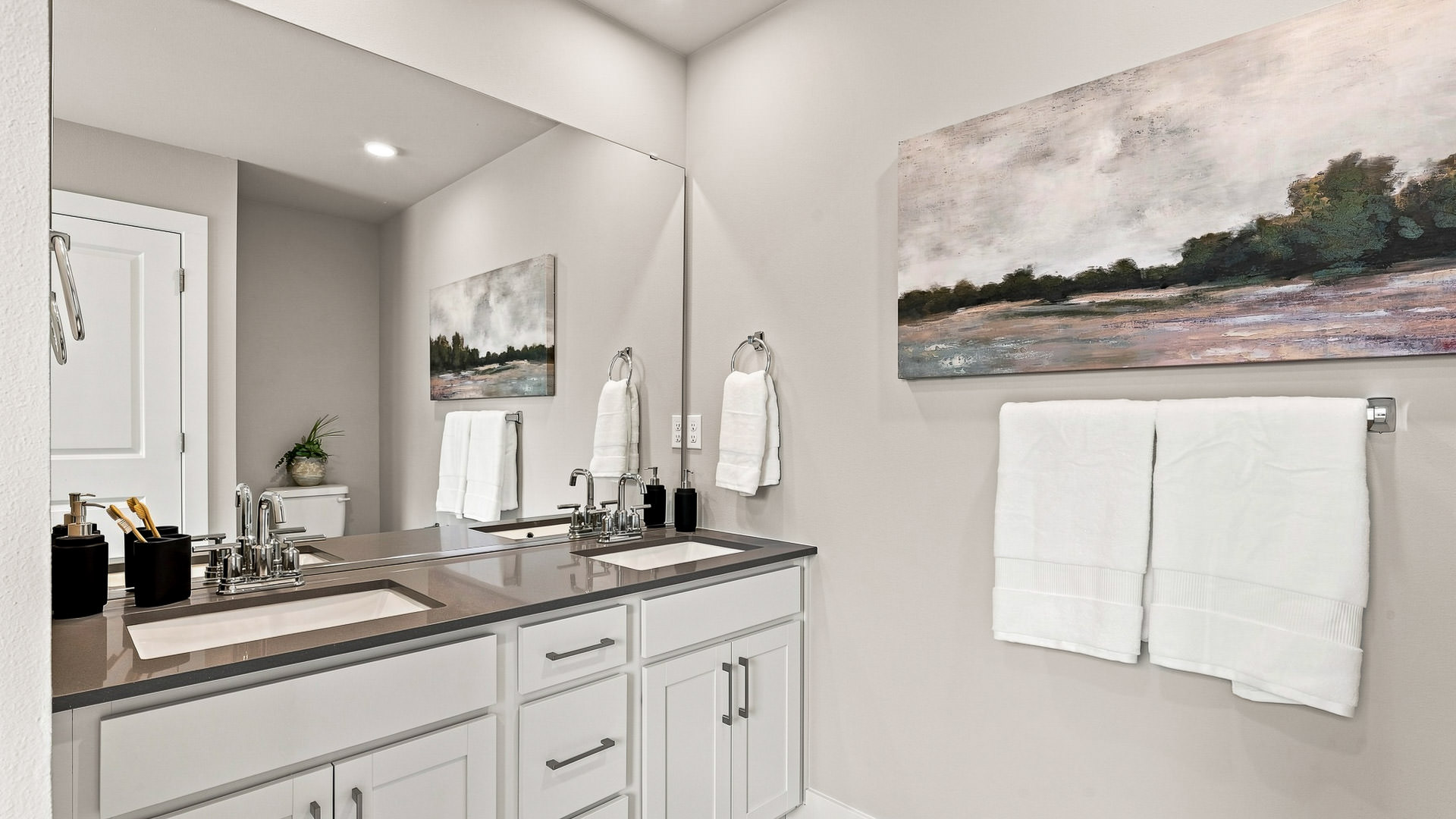 Bathroom with a double-sink vanity, gray countertop, large wall mirror, white cabinetry, and towel rack.
