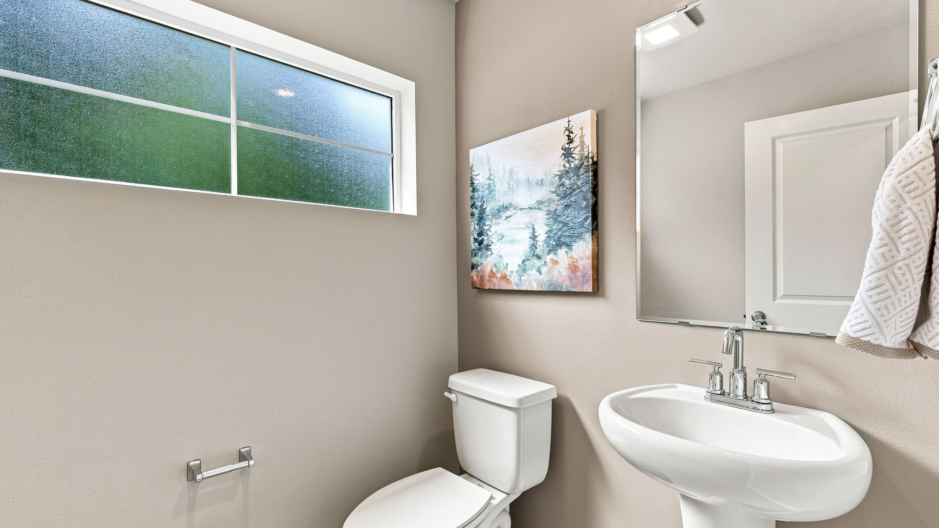 Main floor powder room with a white pedestal sink, chrome fixtures, rectangular mirror, toilet, frosted window, and forest artwork.