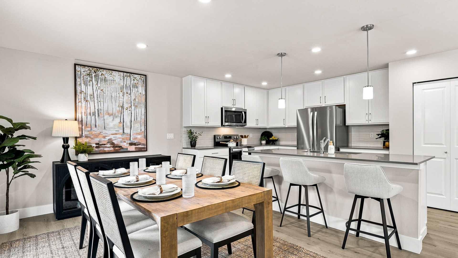 Contemporary dining area with a wooden table set for six adjacent to a bright white kitchen with stainless steel appliances and island seating.