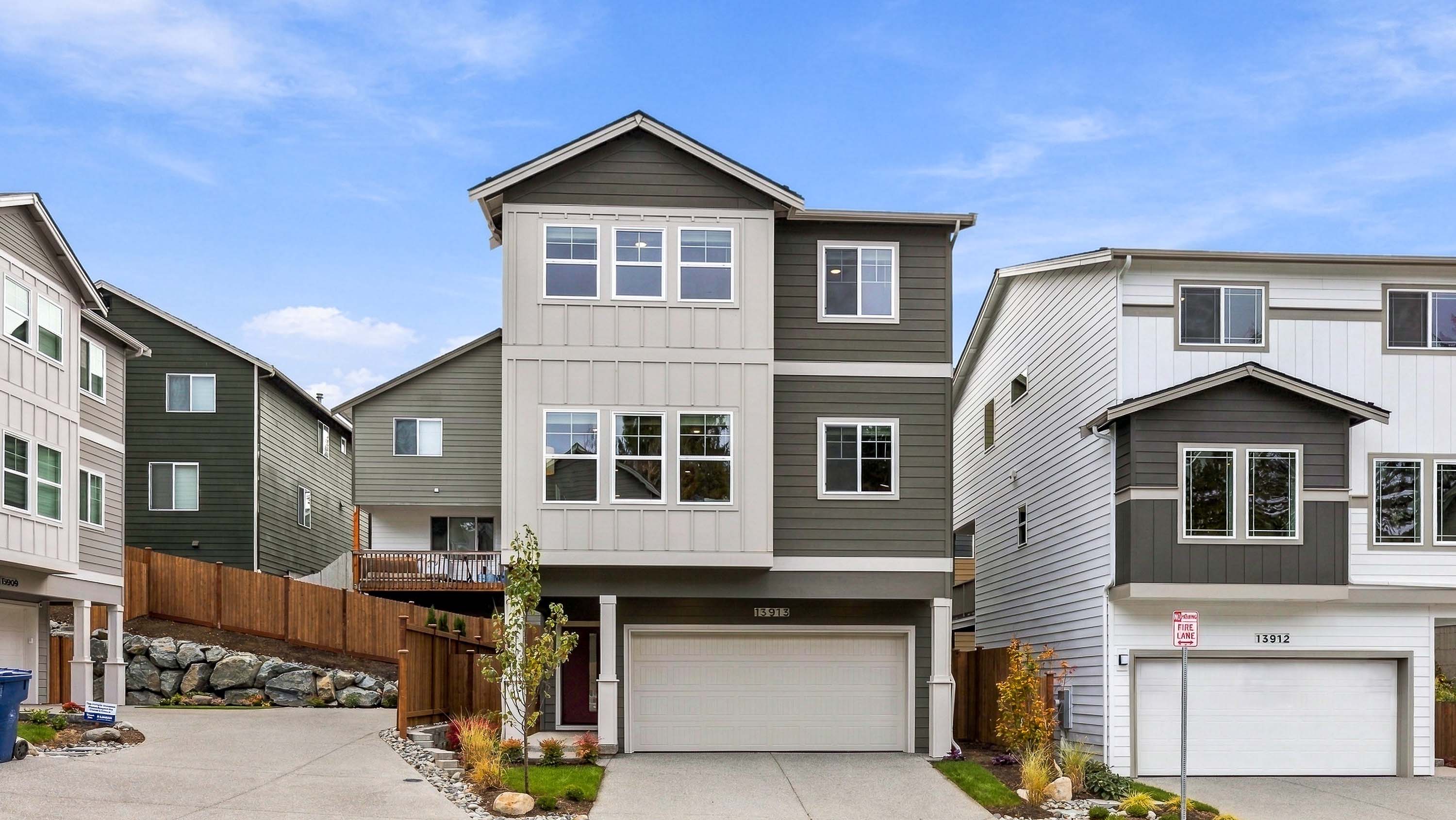 Three-story contemporary home with gray and white siding, multiple front windows, an attached two-car garage, driveway, and landscaped entry.