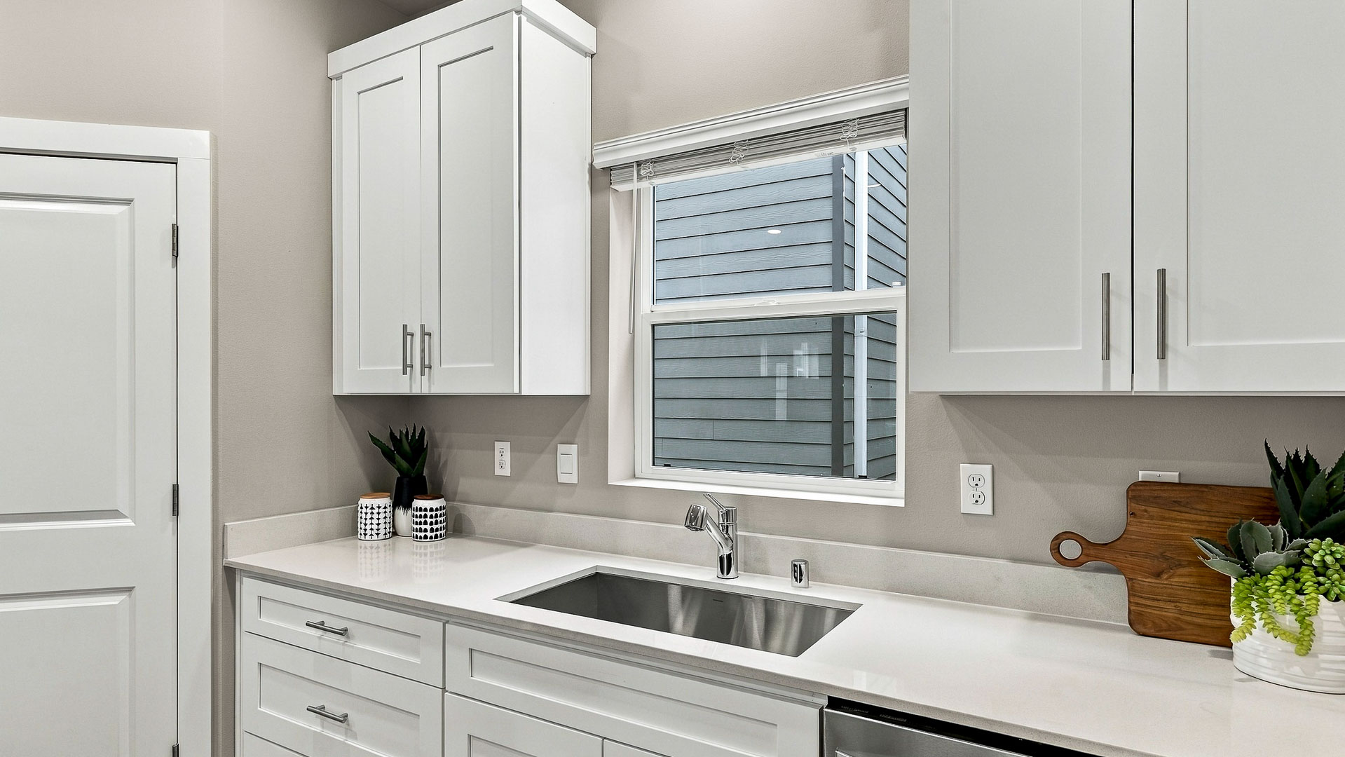 Kitchen sink beneath a window, surrounded by white shaker cabinets, quartz countertops, stainless steel fixtures, plants, and a wood cutting board.