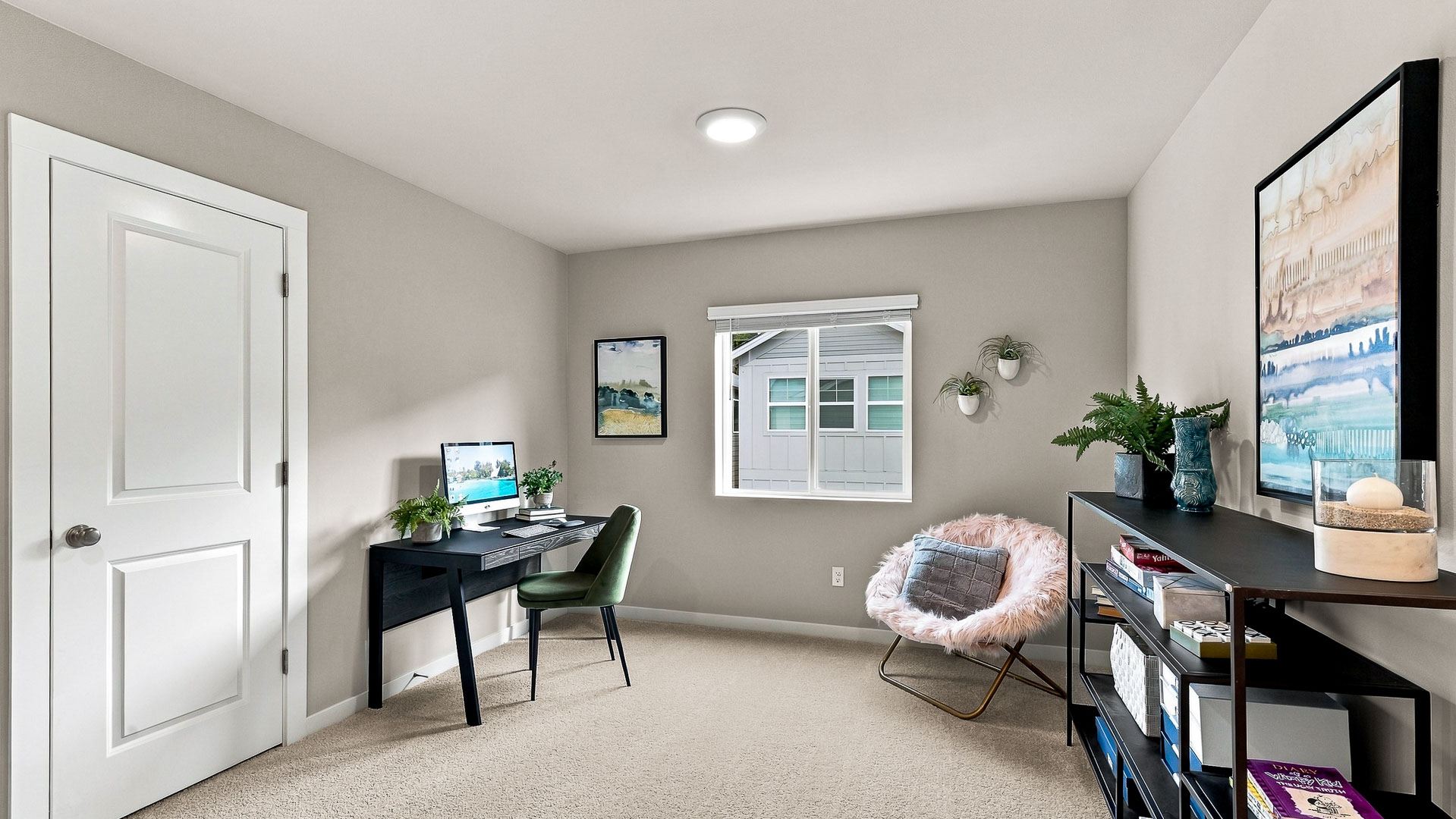 Carpeted home office with a black desk and computer, green desk chair, pink accent chair, open shelving, plants, and framed artwork.