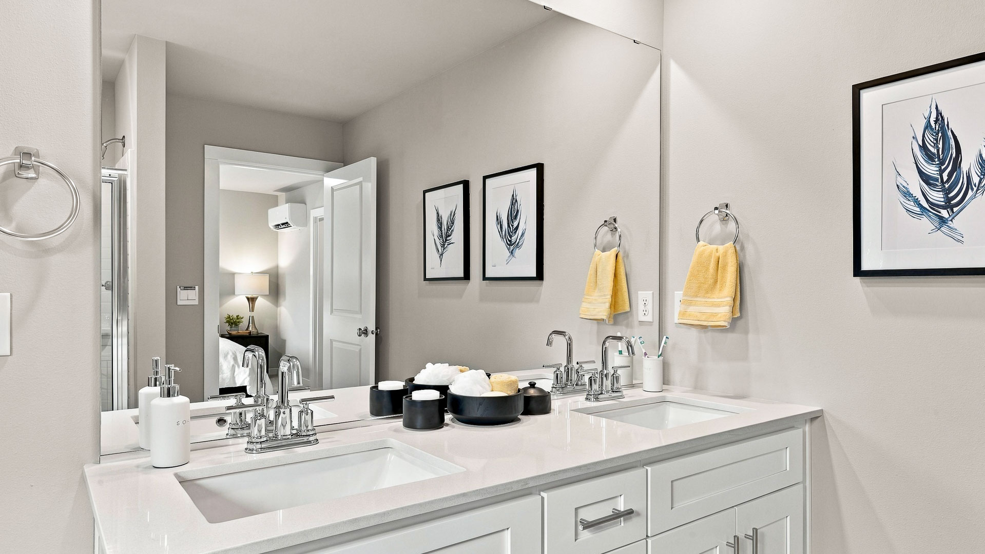 Bathroom with a white double-sink vanity, quartz countertop, large wall mirror, chrome fixtures, yellow hand towels, and framed feather artwork.