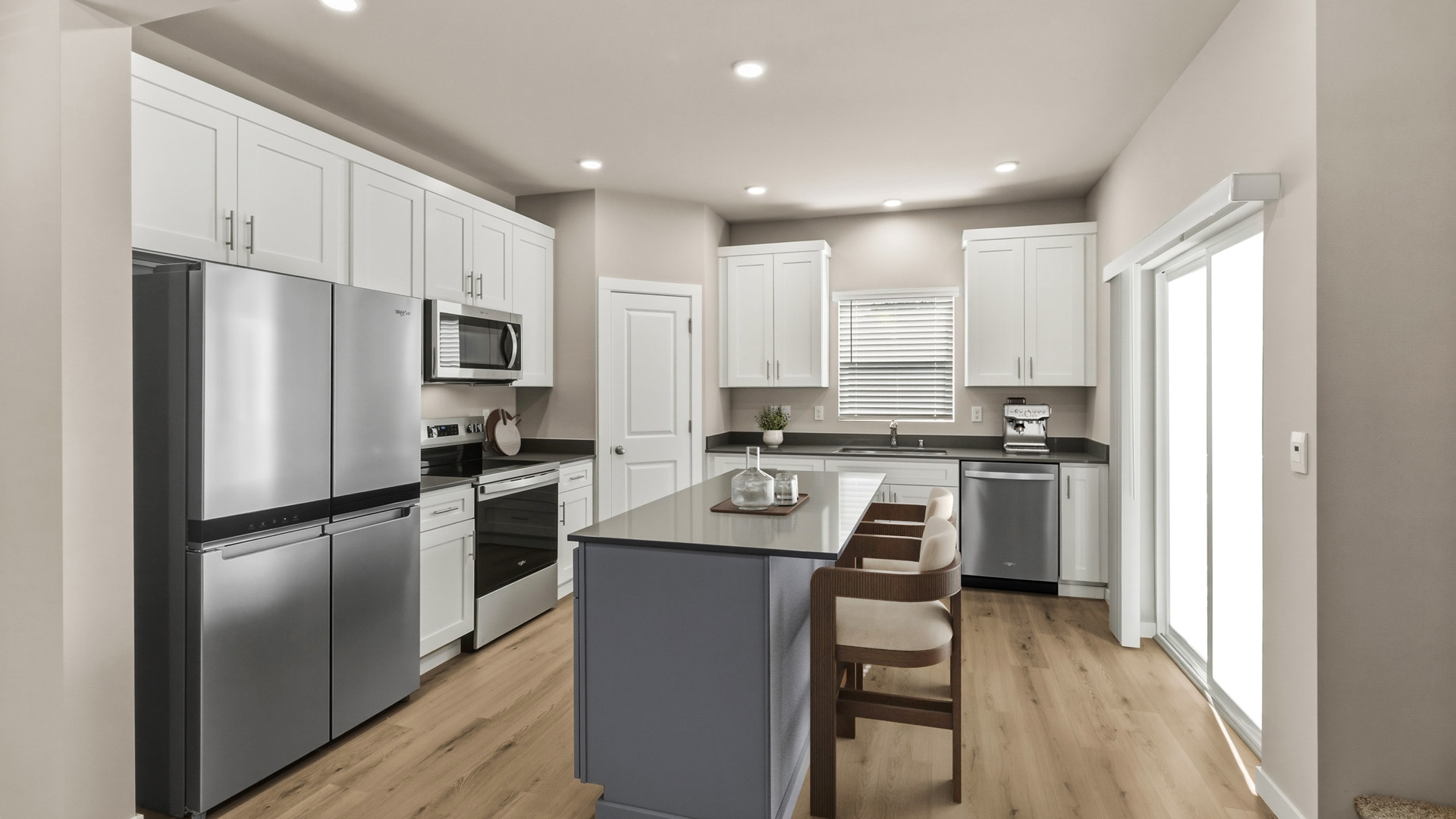 Bright kitchen viewed toward a gray center island, with white cabinetry, stainless steel appliances, two counter stools, and sliding glass doors.