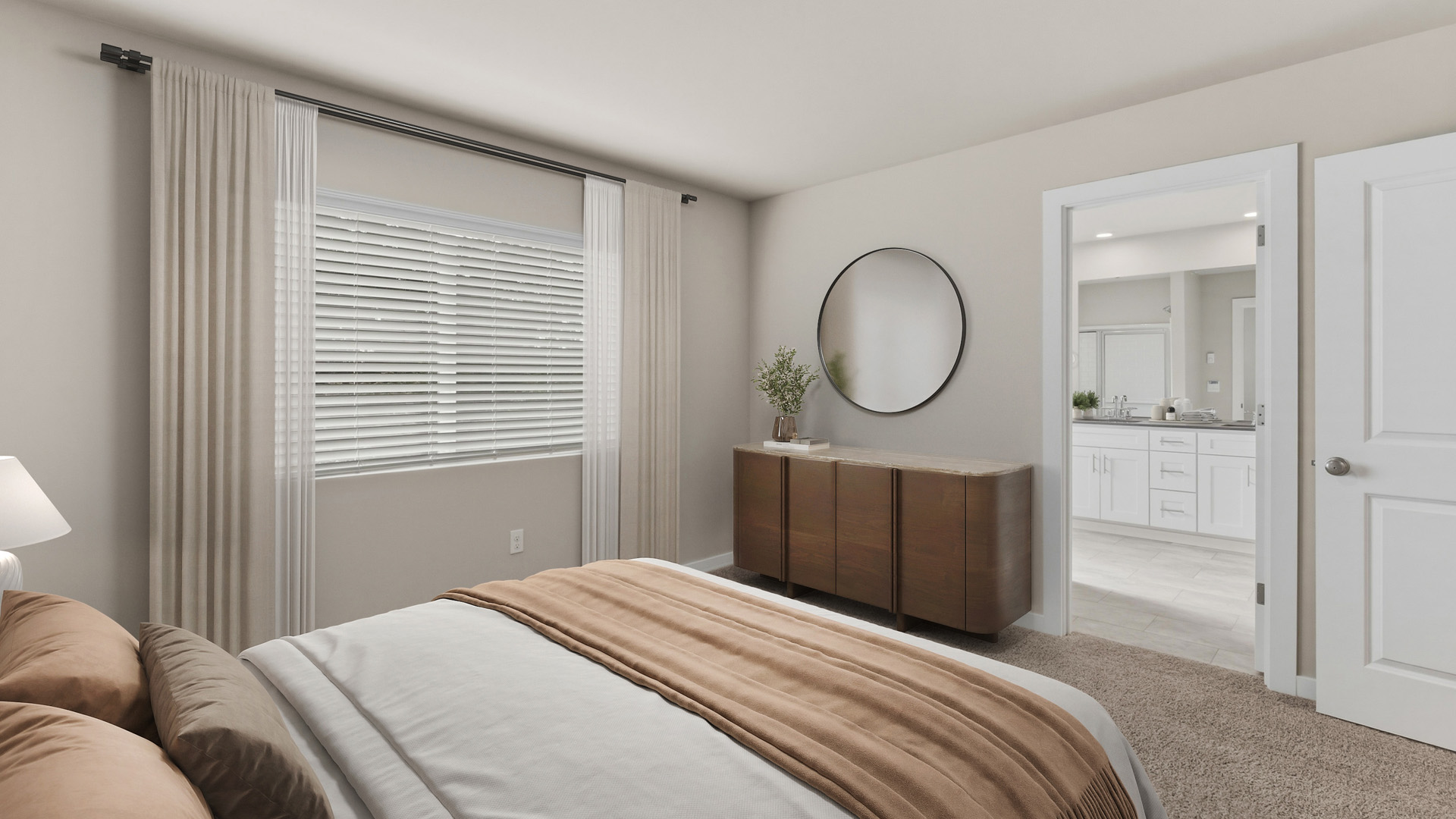 Bedroom view across the foot of a cream bed toward a wood dresser and round mirror, with a large window and an open doorway to a white bathroom vanity.