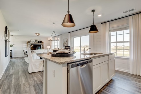 Center island in an open-concept kitchen with white, shaker-style cabinets, statement pendant lights, an undermount sink, and a stainless-steel dishwasher.