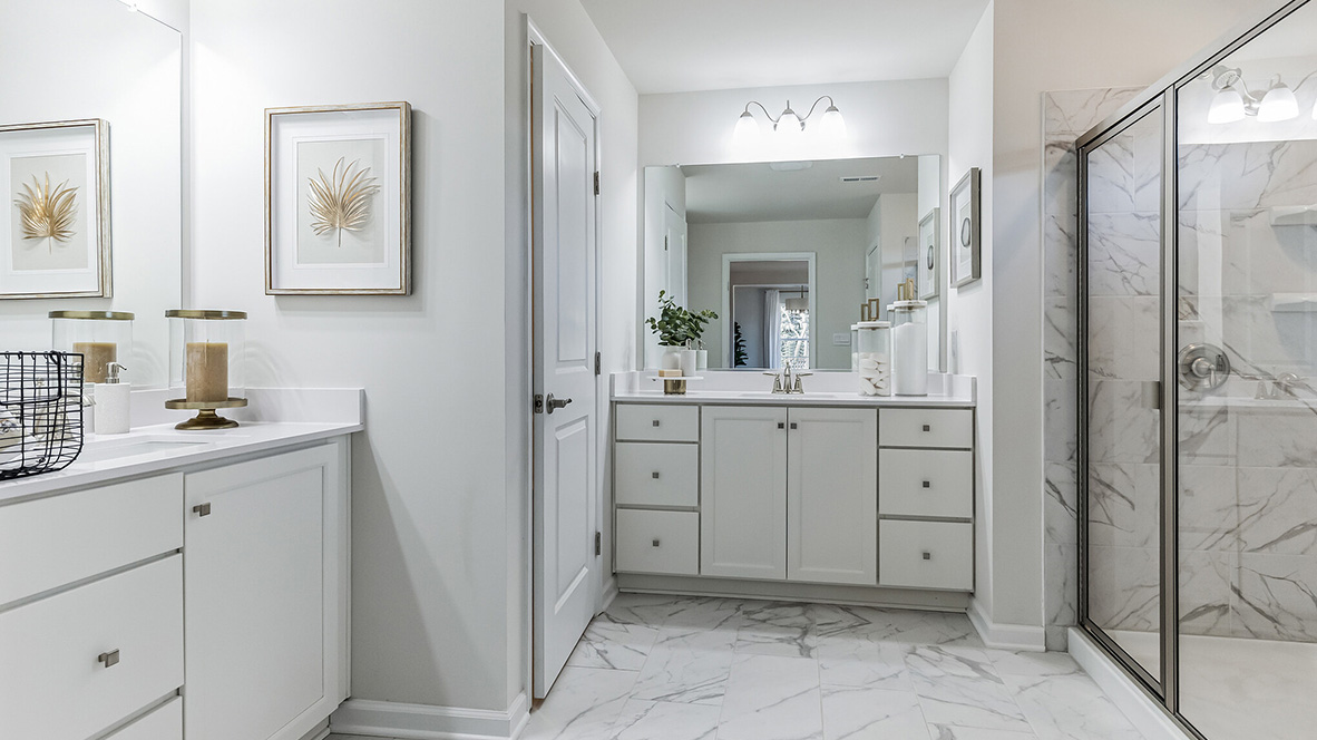 Owners suite connecting bathroom with white and gray marble tile and two separate single sink white vanities. Glass shower door with separate room to the toilet.