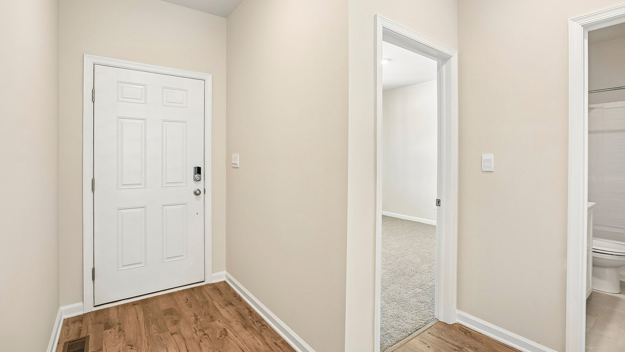Entry foyer with wood-look flooring, white front door, and adjacent hallway leading toward a carpeted bedroom and bathroom.