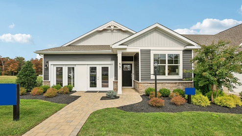 An aerial exterior photograph of a single-story home with gray siding above a stone water table, board-and-batten accents in the gables, and a sales center storefront in place of the two-car garage.