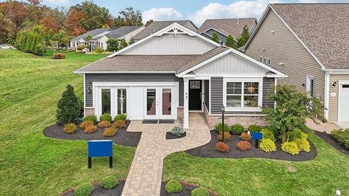 An aerial exterior photograph of a single-story home with gray siding above a stone water table, board-and-batten accents in the gables, and a sales center storefront in place of the two-car garage.