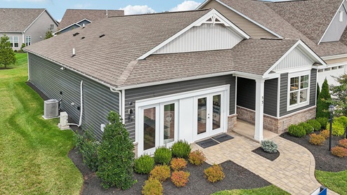 An aerial exterior photograph of a single-story home with gray siding above a stone water table, board-and-batten accents in the gables, and a sales center storefront in place of the two-car garage.