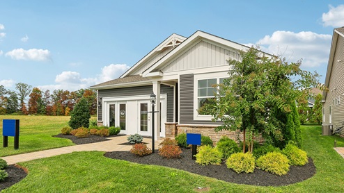 An aerial exterior photograph of a single-story home with gray siding above a stone water table, board-and-batten accents in the gables, and a sales center storefront in place of the two-car garage.