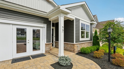 An aerial exterior photograph of a single-story home with gray siding above a stone water table, board-and-batten accents in the gables, and a sales center storefront in place of the two-car garage.