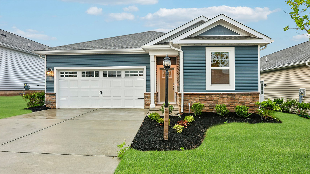 An aerial exterior photograph of a single-story home with dark blue siding above a stone water table, shake accents in the gables, and a white two-car garage with door glass and carriage style hardware