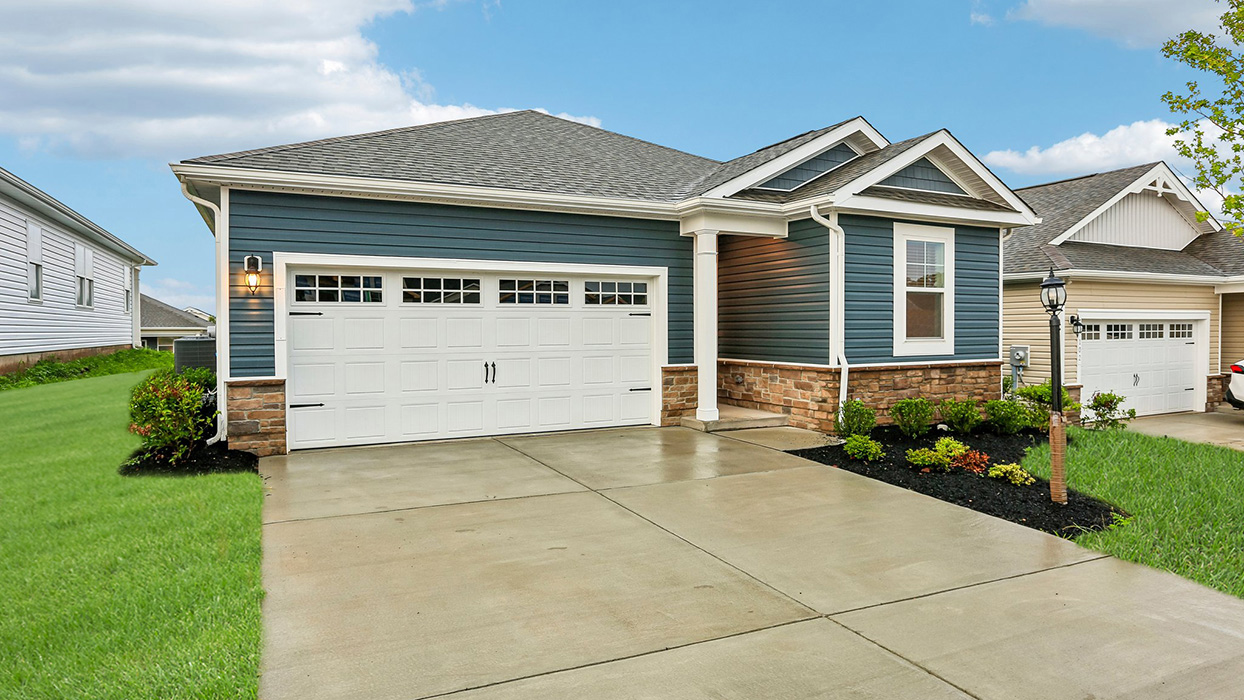 An aerial exterior photograph of a single-story home with dark blue siding above a stone water table, shake accents in the gables, and a white two-car garage with door glass and carriage style hardware