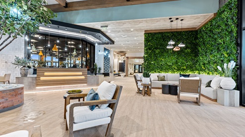 Sitting area with brick-look wallpaper on a focal wall, seating groups of upholstered bench seats and accent chairs with small tables, a tree covered in faux greenery above a banquette, and a tree in a planter.