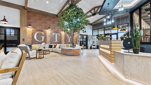 Sitting area with brick-look wallpaper on a focal wall, seating groups of upholstered bench seats and accent chairs with small tables, large wall art, and a tree in a planter.