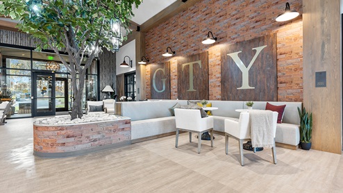 Sitting area with brick-look wallpaper on a focal wall, seating groups of upholstered bench seats and accent chairs with small tables, large wall art, and a tree in a planter.