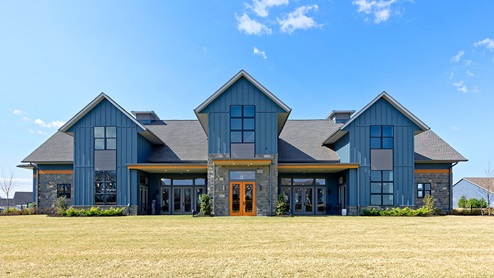 Exterior of the community clubhouse with blue board-and-batten siding, stacked stone accents, modern black-trim windows, and orange doors and trim.