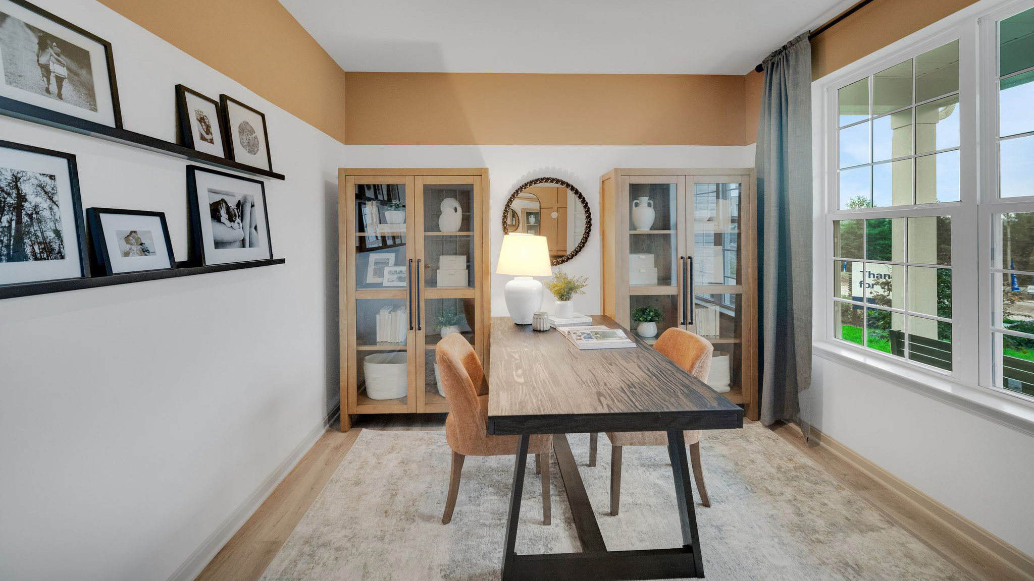Secondary bedroom set up as an office with a desk with chairs on each side, two glass-fronted bookshelves, and picture rails displaying family photos on the wall opposite of the window.