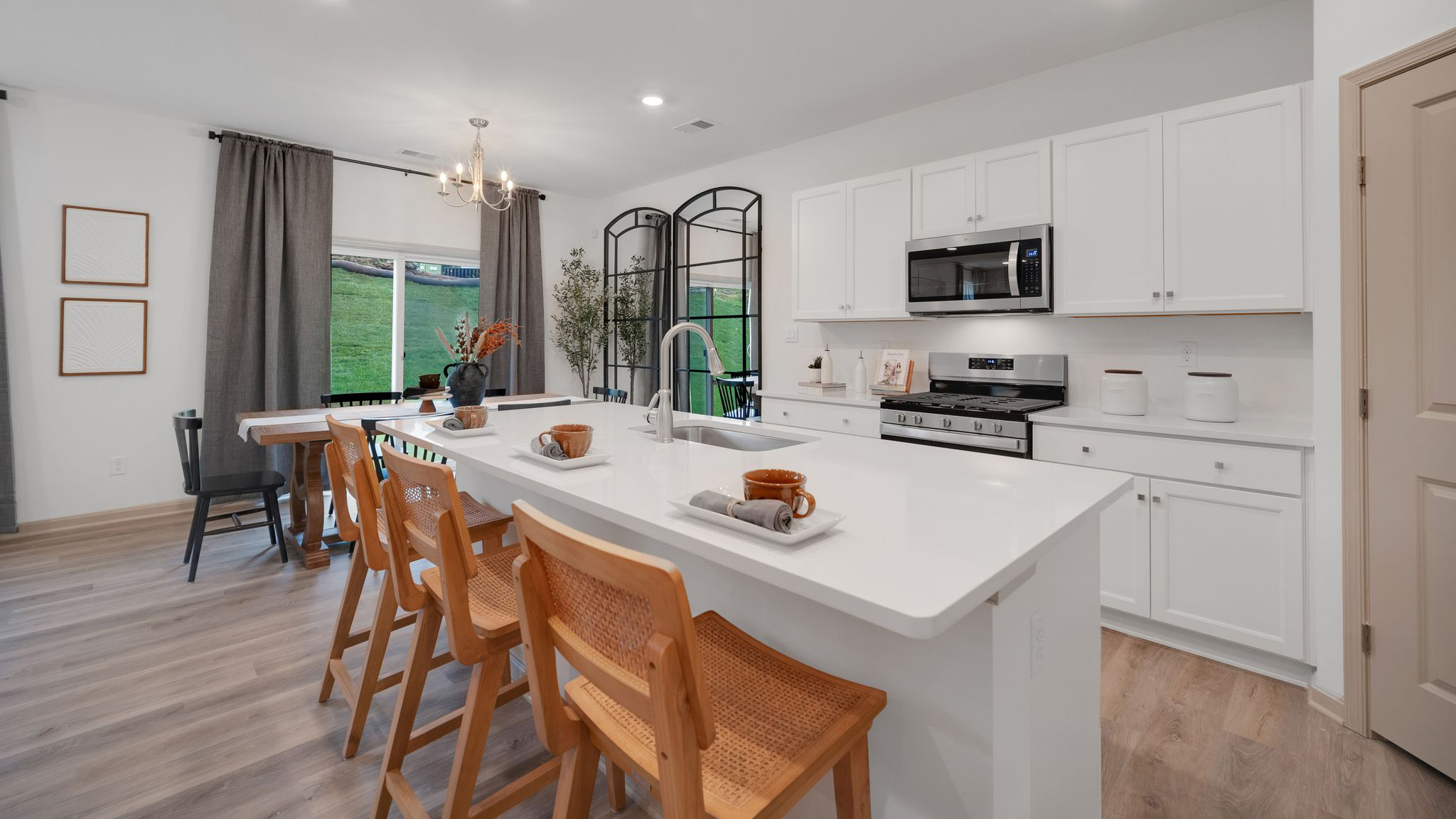 A spacious kitchen with white, shaker-style cabinets, quartz countertops, a large center island with undermount sink and seating for three, and a stainless-steel appliance package that flows into an adjoining dining area that boasts a wooden table surrounded by six chairs.
