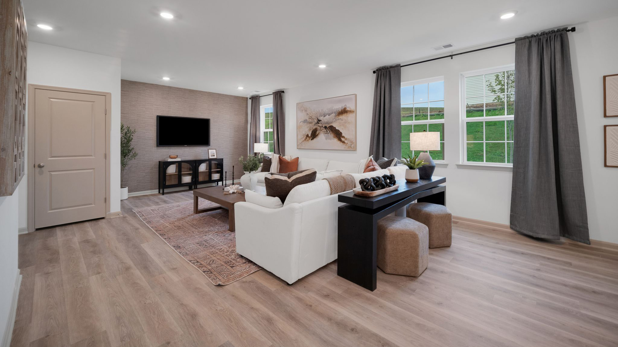 Living room with a large sectional sofa and coffee table arranged facing a wall-mounted tv above a storage console, all in front of a wall boasting three windows overlooking the back yard.