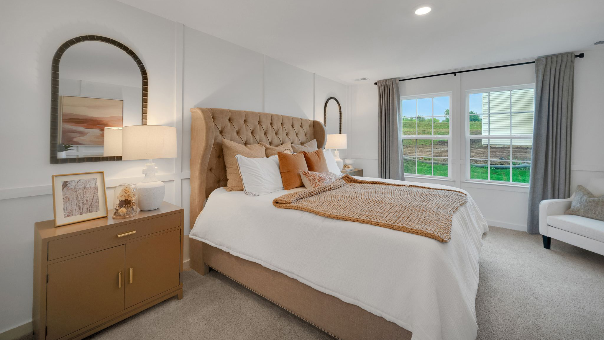Primary bedroom with a large bed with upholstered headboard flanked by two nightstands with an accent chair in the corner next to the double window.