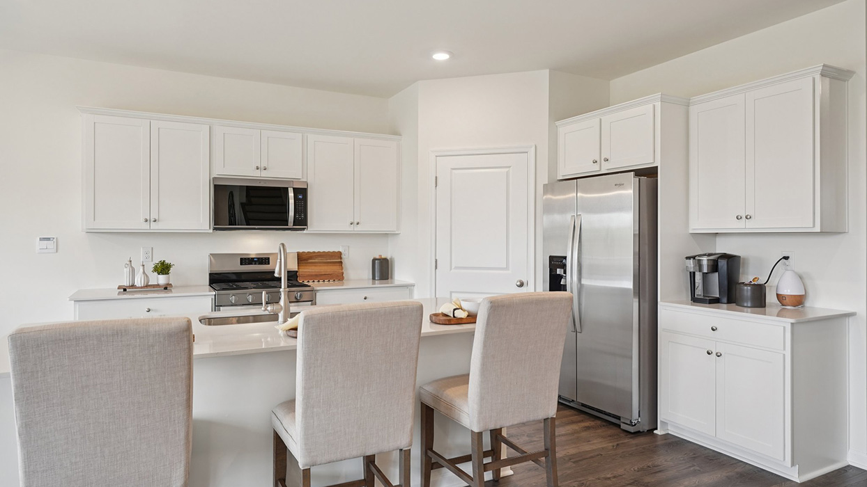 A spacious kitchen with white, shaker-style cabinets, quartz countertops, a large center island with undermount sink and seating for three, and a stainless-steel appliance package overlooking the adjoining dining space and living room.